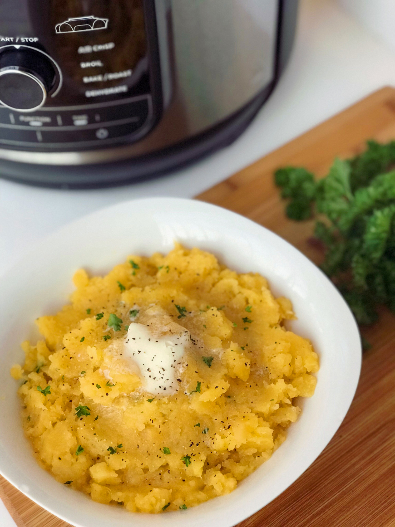 a white plate of mashed rutabaga topped with melted butter and fresh chives sitting in front of the pressure cooker that was used to cook them