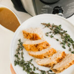 a hand holding a plate of sliced herbed turkey breast with a pressure cooker and gluten free gravy in the background